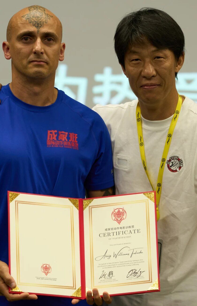Aang Willians segura o certificado de participação do JCST Action Film Camp 2025, em Pequim, ao lado de um instrutor. Foto de 2025, publicada em 2026, do Instagram dele.