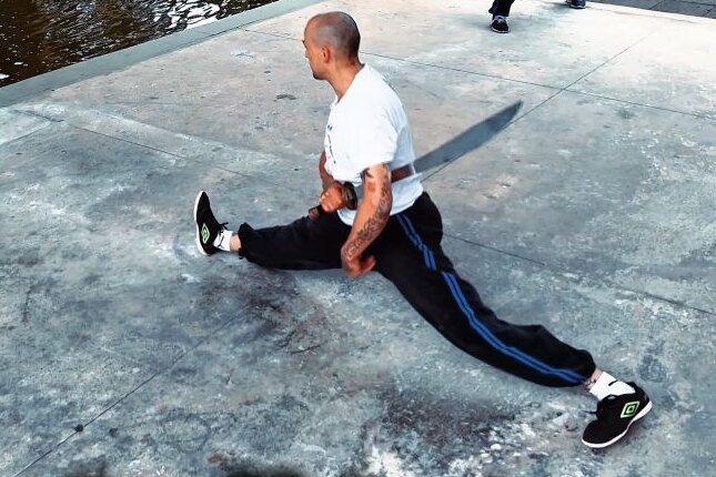 Aang Willians em espacate no chão de um parque, com um facão na mão. 2025 (vídeo de @taiada_drone), do Instagram dele.