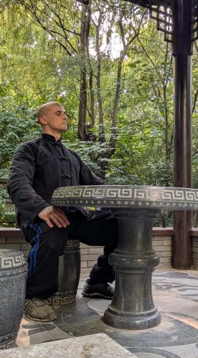 Aang Willians de jaqueta chinesa preta, sentado de perfil a uma mesa de pedra, num pavilhão de jardim em Pequim. 2025, do Instagram dele.