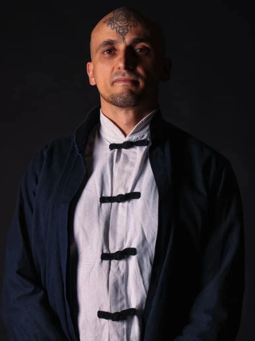 Aang Willians de jaqueta chinesa escura sobre camisa branca com botões de alamar, diante de um fundo escuro. Retrato publicado em 2022 na Dublagem Wiki.