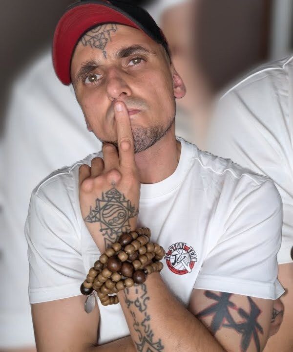 Aang Willians de boné, com o dedo nos lábios, contas de madeira no pulso e tatuagens nas mãos e nos braços. 6 de maio de 2026, do canal dele no YouTube.