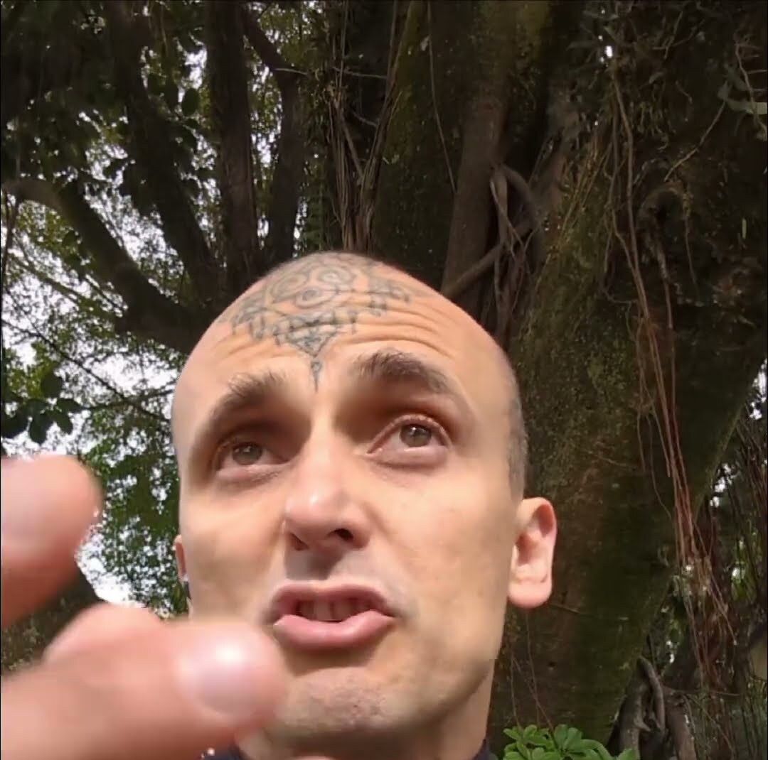 Aang Willians de jaqueta chinesa preta fala para a câmera debaixo das árvores. 19 de junho de 2025, do canal dele no YouTube.
